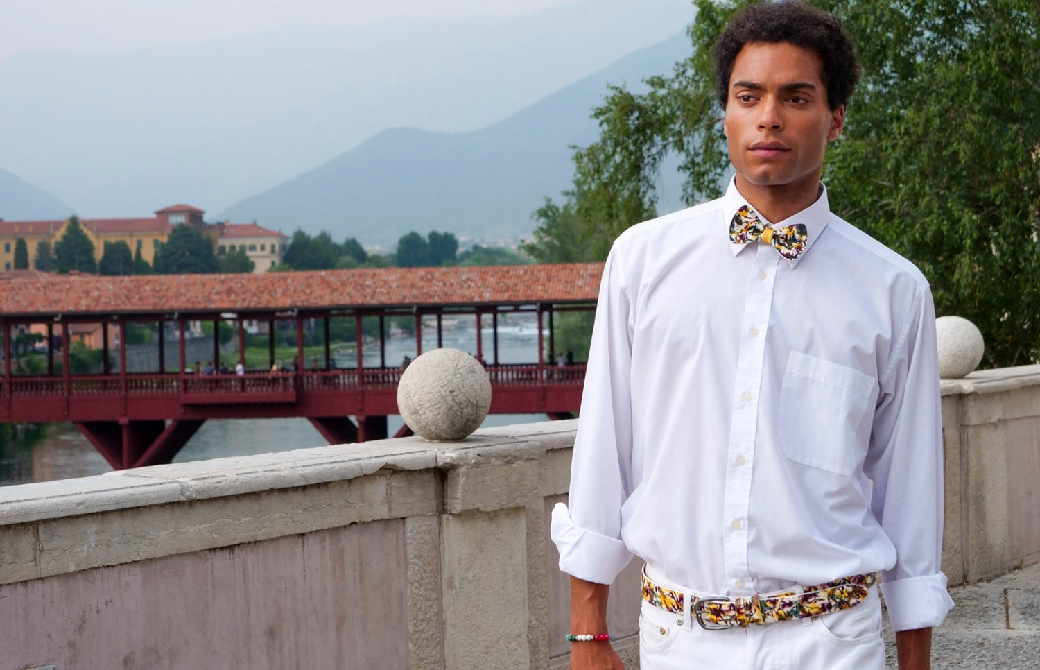 model in white with a yellow black and white belt and bow tie and Ponte Degli Alpini bridge on the background
